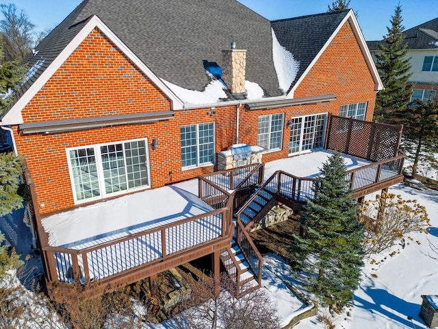 snow covered house featuring a deck, roof with shingles, brick siding, and a chimney