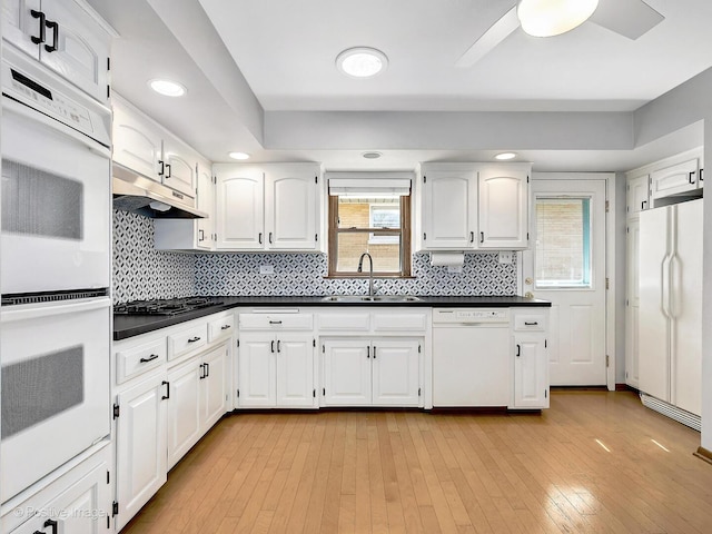 kitchen with white appliances, a sink, under cabinet range hood, dark countertops, and light wood-type flooring