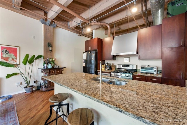 kitchen with black refrigerator, stainless steel range with gas stovetop, light stone counters, light hardwood / wood-style floors, and wall chimney range hood