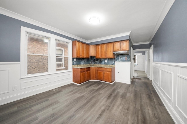 kitchen with dark wood-style floors, a wainscoted wall, brown cabinetry, ornamental molding, and under cabinet range hood