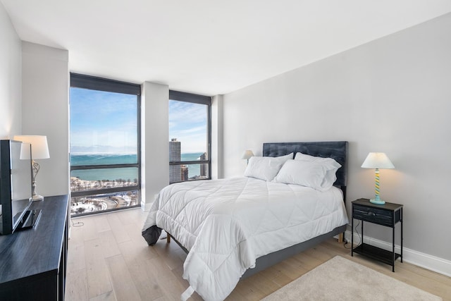 bedroom featuring light wood-type flooring, floor to ceiling windows, a water view, and baseboards