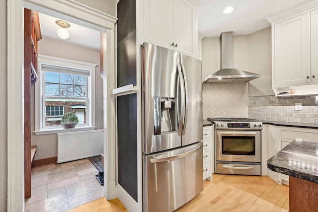 kitchen featuring white cabinetry, appliances with stainless steel finishes, wall chimney range hood, backsplash, and radiator heating unit