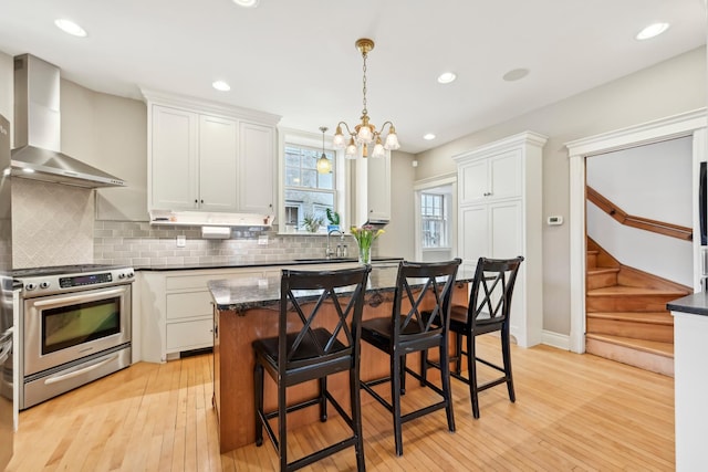 kitchen featuring light wood finished floors, stainless steel range with gas stovetop, wall chimney range hood, white cabinetry, and backsplash