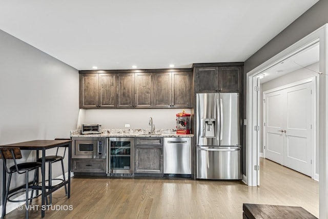 kitchen featuring dark brown cabinetry, light wood finished floors, wine cooler, light stone counters, and stainless steel appliances