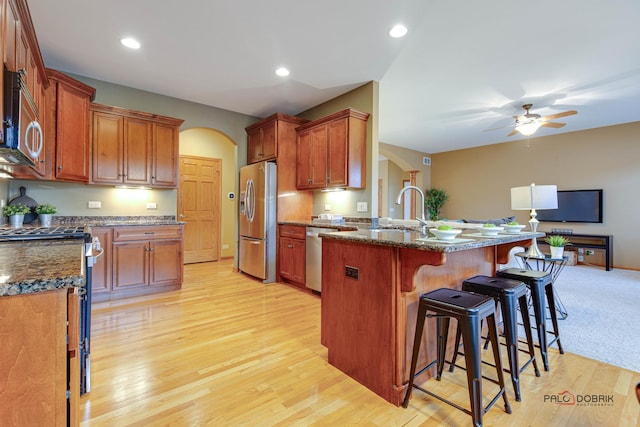 kitchen with a kitchen bar, dark stone counters, ceiling fan, light hardwood / wood-style floors, and stainless steel appliances