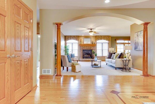 living room with a brick fireplace, light wood-type flooring, ceiling fan, and ornate columns