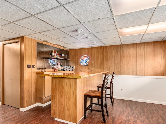 bar featuring dark wood-type flooring, a drop ceiling, a bar, wood walls, and baseboards