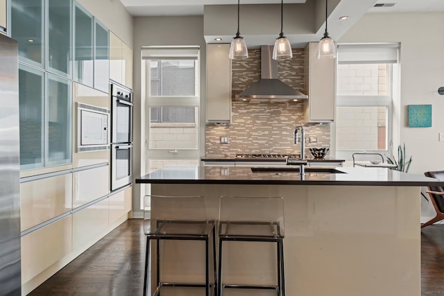 kitchen with pendant lighting, wall chimney range hood, sink, appliances with stainless steel finishes, and a kitchen island with sink