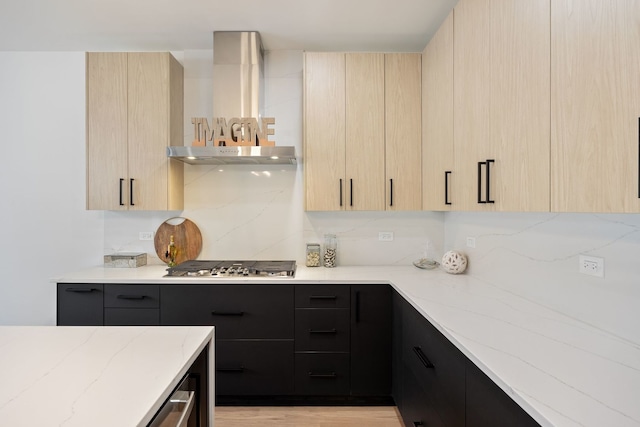 kitchen with light stone counters, stainless steel gas stovetop, light brown cabinetry, wall chimney range hood, and dark cabinetry