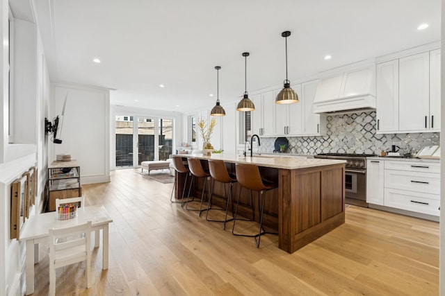 kitchen featuring white cabinets, an island with sink, high end range, custom exhaust hood, and pendant lighting