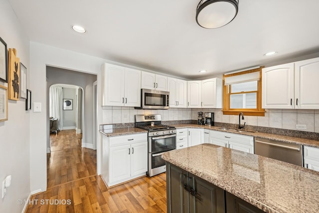 kitchen with arched walkways, stainless steel appliances, dark stone counters, a sink, and light wood-type flooring