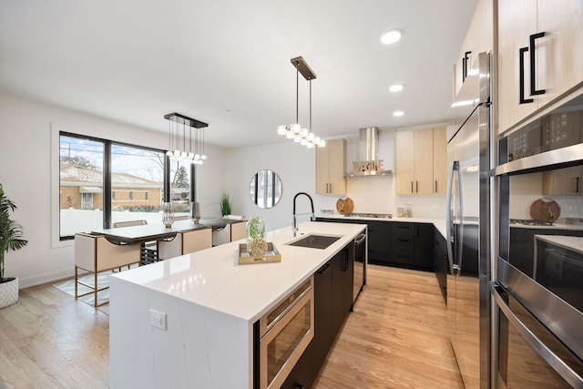 kitchen featuring a kitchen island with sink, stainless steel appliances, a sink, light countertops, and wall chimney range hood