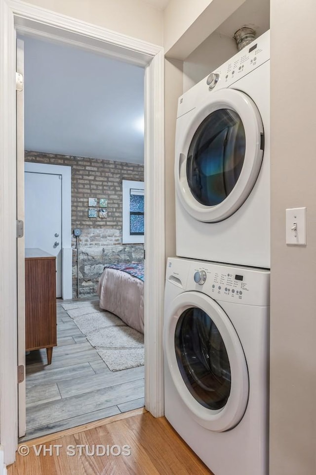 laundry room featuring brick wall, laundry area, wood finished floors, and stacked washer and clothes dryer