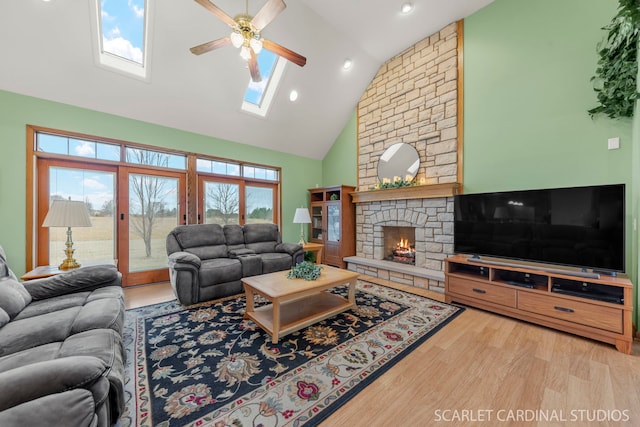 living room featuring a stone fireplace, high vaulted ceiling, a skylight, ceiling fan, and light hardwood / wood-style flooring