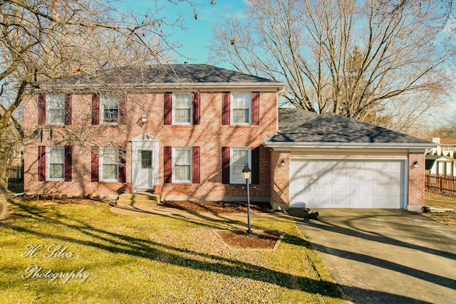 colonial home with a garage, driveway, roof with shingles, fence, and brick siding