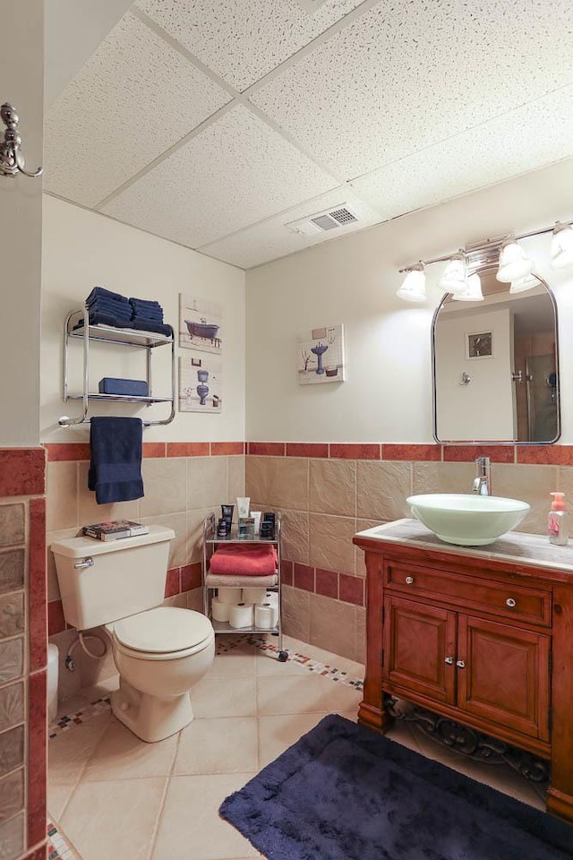 bathroom with a paneled ceiling, tile walls, tile patterned flooring, vanity, and toilet