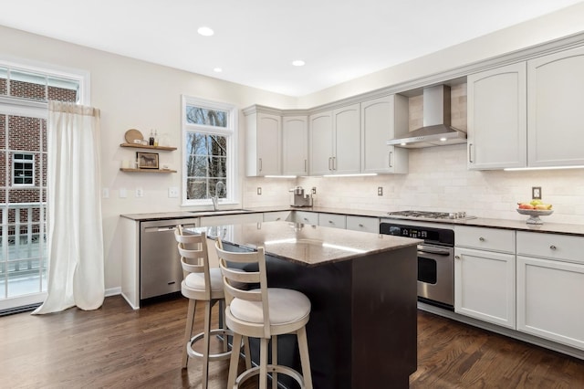 kitchen featuring sink, appliances with stainless steel finishes, a kitchen island, light stone countertops, and wall chimney range hood