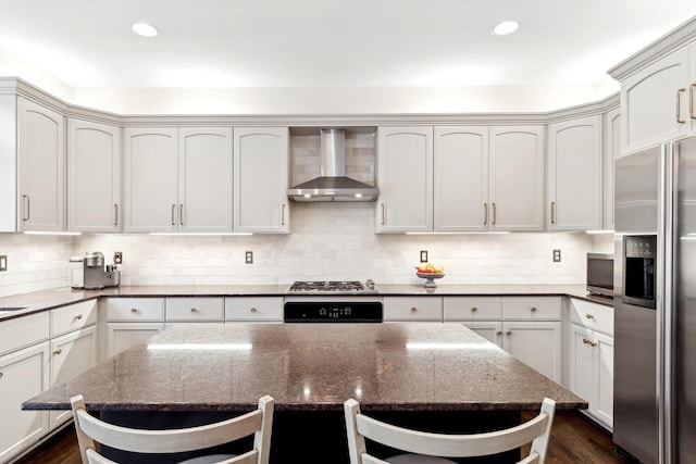 kitchen with wall chimney range hood, stainless steel appliances, dark hardwood / wood-style flooring, decorative backsplash, and dark stone counters