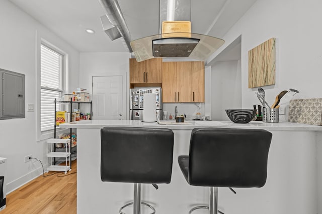 kitchen with electric panel, island exhaust hood, light wood-type flooring, stainless steel fridge, and a breakfast bar area