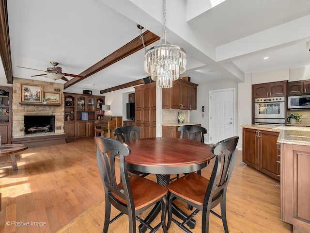 dining room with light wood-style floors, a fireplace, beamed ceiling, and ceiling fan with notable chandelier