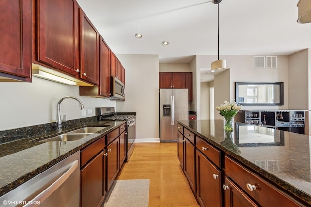 kitchen with sink, appliances with stainless steel finishes, dark stone countertops, light hardwood / wood-style floors, and decorative light fixtures