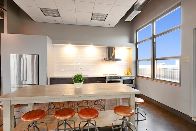 kitchen featuring wall chimney range hood, a paneled ceiling, a breakfast bar, backsplash, and stainless steel appliances