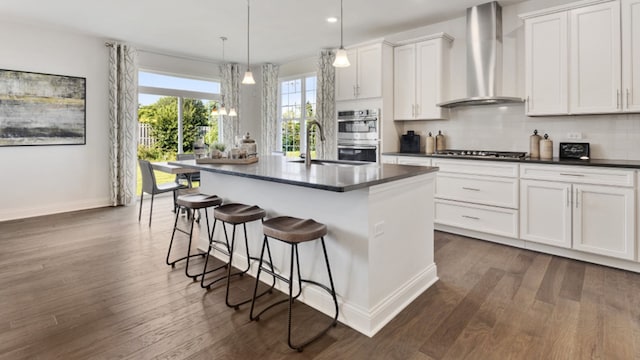 kitchen with wall chimney exhaust hood, sink, decorative light fixtures, a kitchen island with sink, and white cabinets