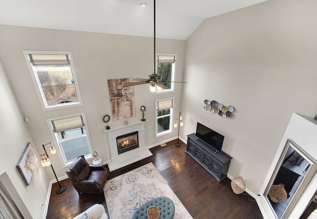 living area featuring dark wood-style floors, a glass covered fireplace, ceiling fan, high vaulted ceiling, and baseboards