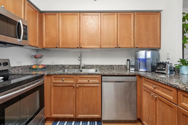 kitchen featuring brown cabinetry, appliances with stainless steel finishes, dark stone counters, and a sink