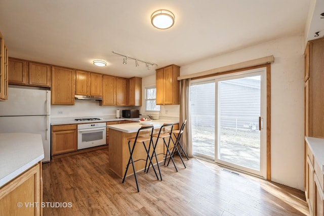 kitchen featuring under cabinet range hood, light countertops, a kitchen breakfast bar, wood finished floors, and white appliances