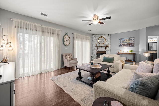 living room with ceiling fan, a stone fireplace, dark hardwood / wood-style floors, and a healthy amount of sunlight