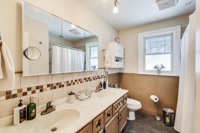 full bath with double vanity, visible vents, toilet, wainscoting, and a sink