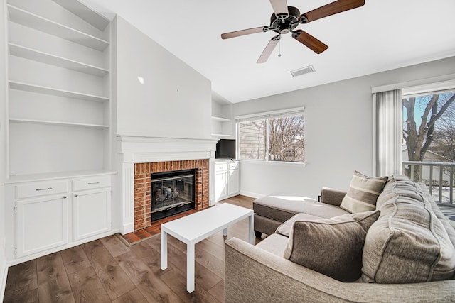 living area featuring built in features, visible vents, a ceiling fan, a brick fireplace, and wood finished floors