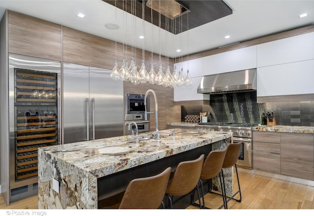 kitchen with white cabinets, a large island with sink, stainless steel appliances, light stone countertops, and wall chimney exhaust hood