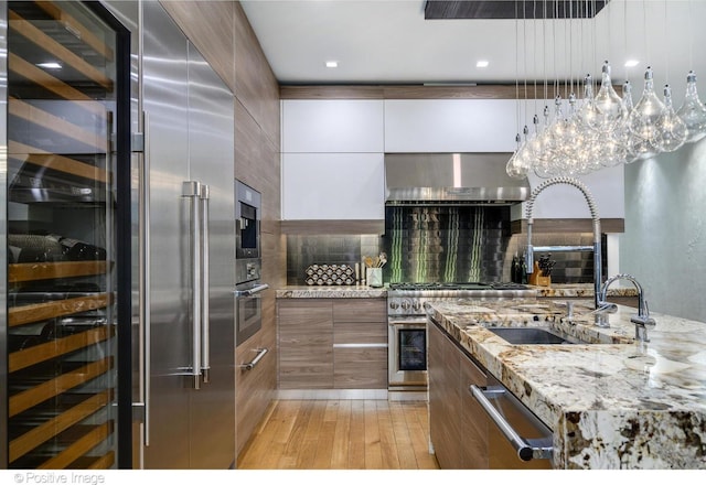 kitchen with decorative backsplash, built in appliances, light hardwood / wood-style floors, light stone counters, and wall chimney range hood