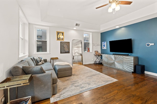 living area with arched walkways, dark wood-style flooring, a raised ceiling, ceiling fan, and baseboards