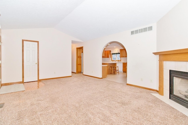 unfurnished living room with arched walkways, a tile fireplace, light colored carpet, visible vents, and vaulted ceiling