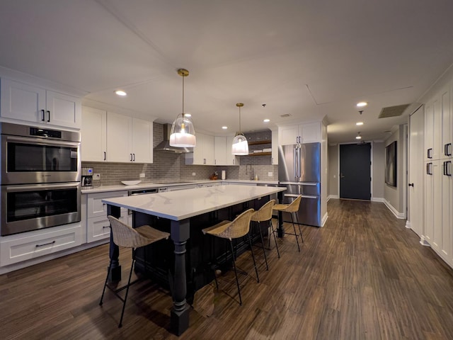 kitchen featuring wall chimney range hood, appliances with stainless steel finishes, hanging light fixtures, white cabinets, and a kitchen island