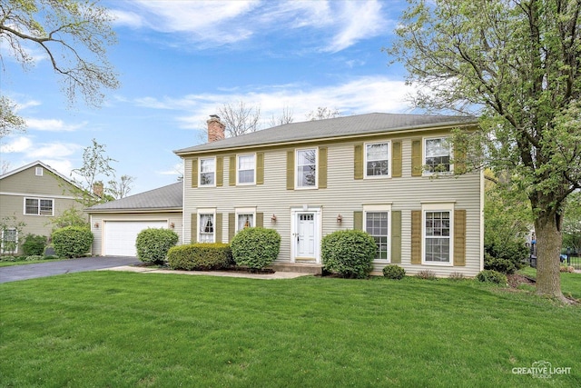 colonial-style house with asphalt driveway, a chimney, a garage, and a front lawn