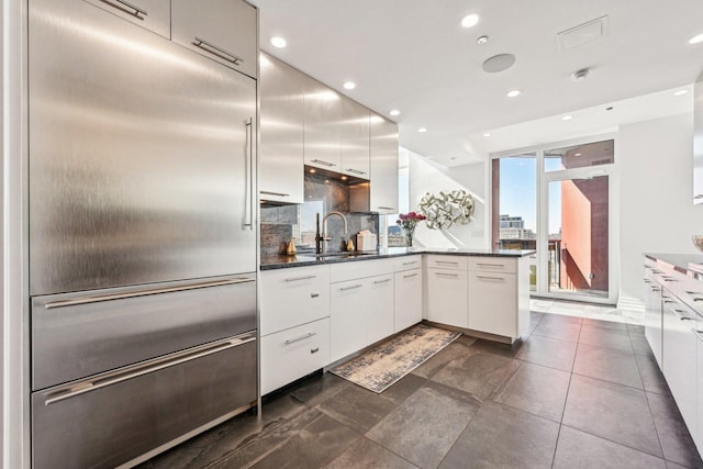 kitchen with a peninsula, a sink, stainless steel built in fridge, white cabinets, and dark countertops