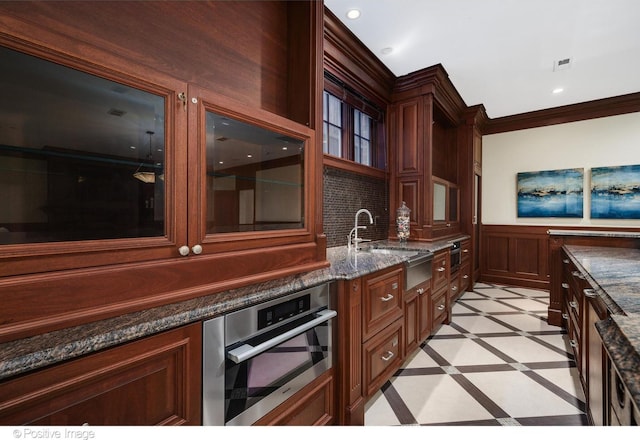 kitchen with a sink, dark stone counters, crown molding, visible vents, and stainless steel oven