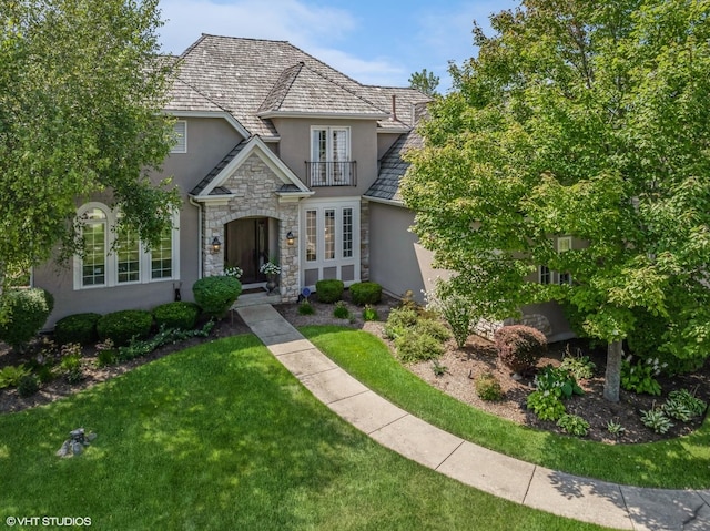 french country home with a front yard, stone siding, a balcony, and stucco siding