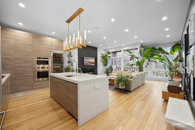 kitchen featuring pendant lighting, a fireplace, stainless steel double oven, a center island with sink, and light hardwood / wood-style flooring