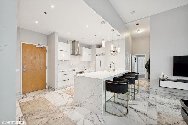 kitchen with sink, white cabinetry, decorative light fixtures, an island with sink, and wall chimney range hood