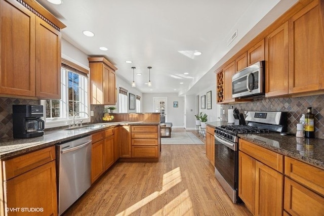 kitchen with stainless steel appliances, a peninsula, light wood-style floors, brown cabinets, and dark stone counters