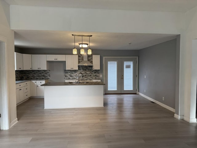 kitchen featuring french doors, a center island with sink, decorative light fixtures, white cabinets, and wall chimney exhaust hood