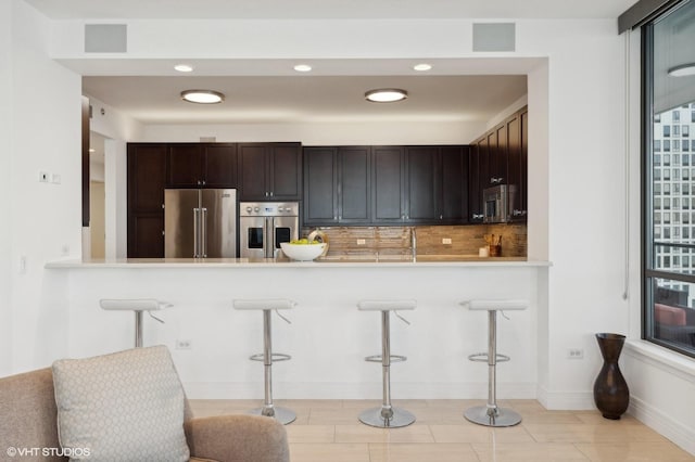 kitchen with tasteful backsplash, dark brown cabinets, appliances with stainless steel finishes, a kitchen breakfast bar, and kitchen peninsula