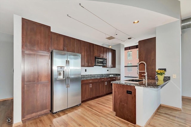 kitchen with stainless steel appliances, a peninsula, a sink, light wood-type flooring, and dark stone counters