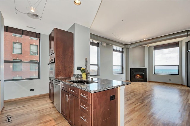 kitchen featuring a warm lit fireplace, light wood finished floors, dark stone counters, stainless steel appliances, and a sink