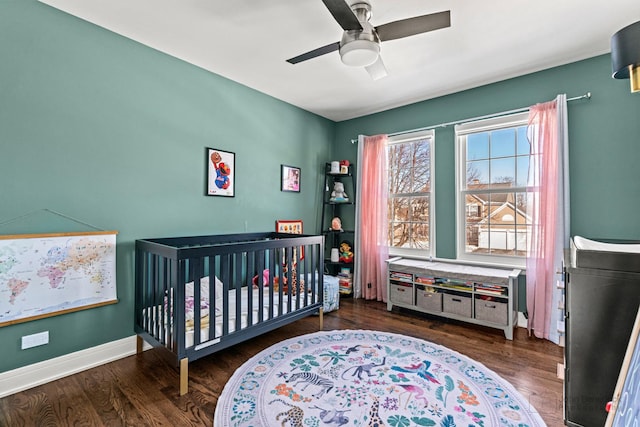 bedroom with a nursery area, dark wood finished floors, baseboards, and ceiling fan